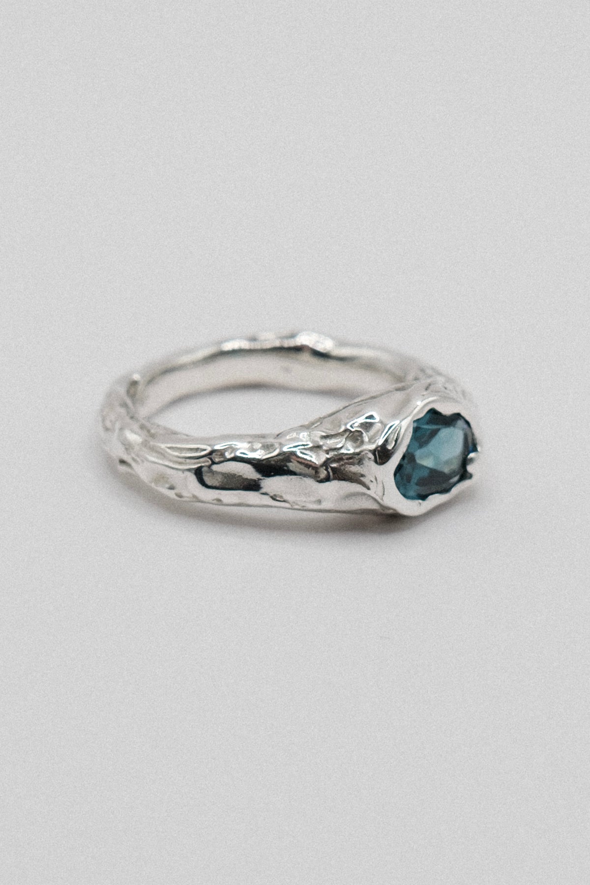 A silver ring with a textured, organic-shaped band and a small, heart-shaped dark blue gemstone, shown on a neutral light gray background.