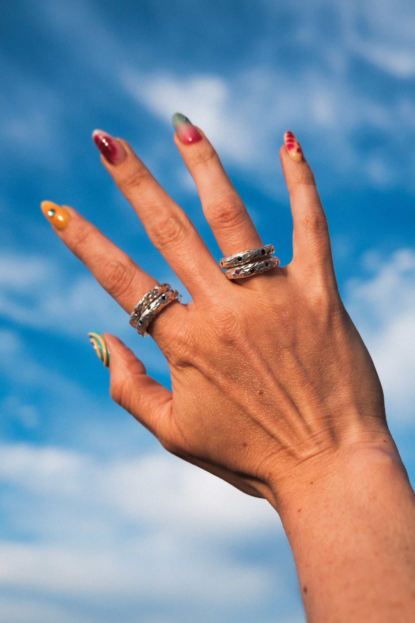 Hand with colorful nail polish and multiple silver rings against a blue sky background