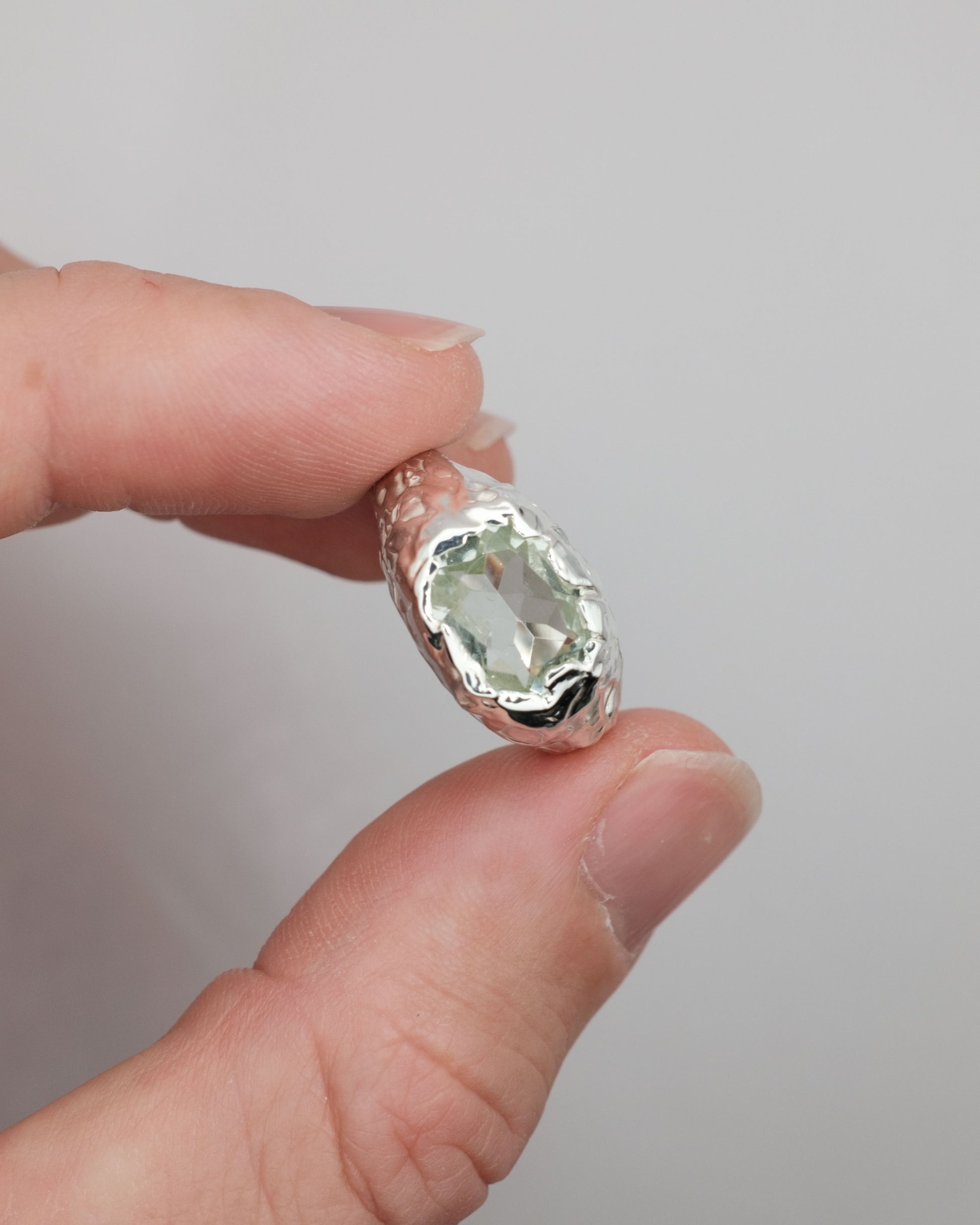 Hand holding a silver ring with a green amethystgemstone against a gray background