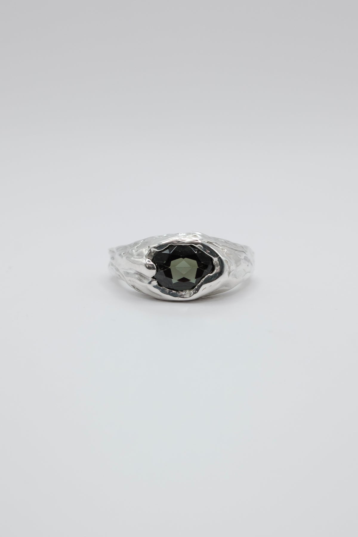 A silver ring with a textured, organic-shaped setting holding a dark green oval gemstone, photographed on a seamless light gray background.