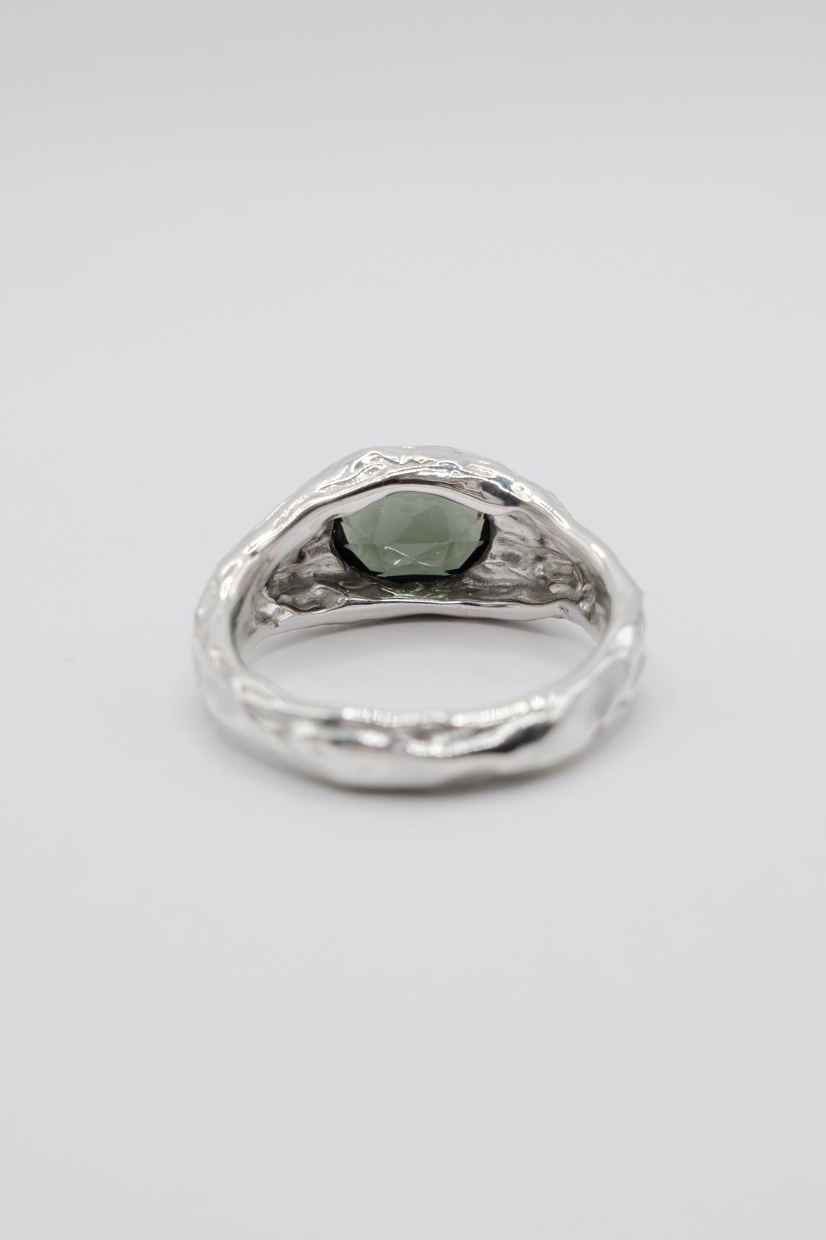 A silver ring with a textured, organic-shaped band and a central oval green gemstone, shown on a neutral light gray background.