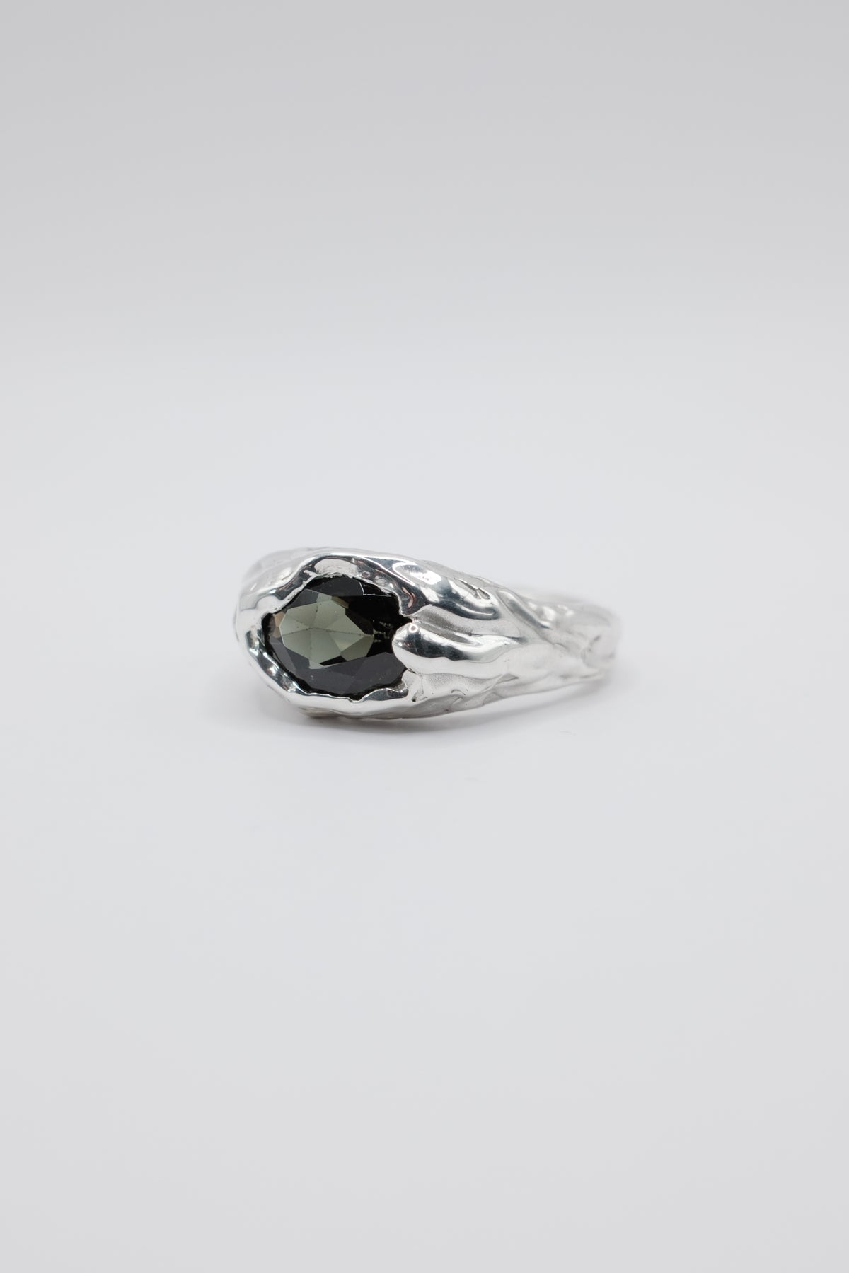 A silver ring featuring a textured, organic-shaped setting holding a dark gray faceted gemstone, photographed on a seamless light gray background.